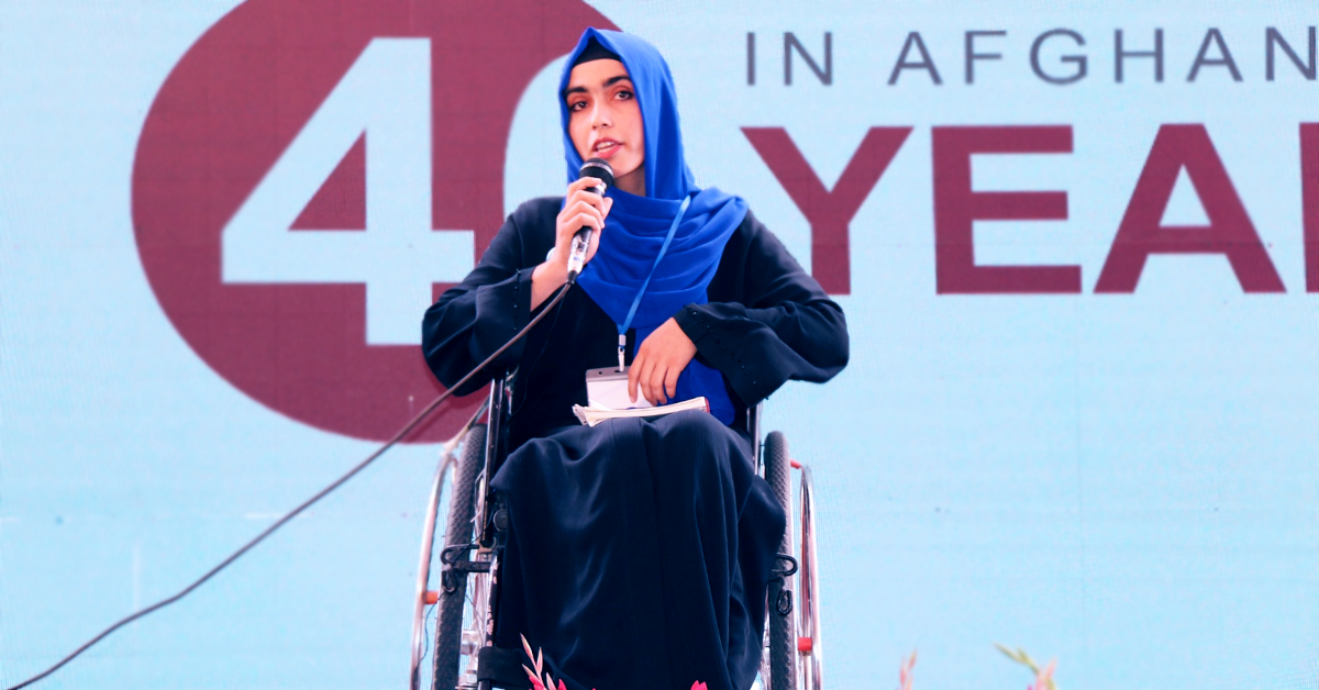 Habiba Noorzad sitter i sin rullstol på en scen och berättar sin historia i en mikrofon.