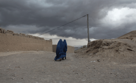 två kvinnor i burka går längs en sandad väg. I bakgrunden syns en stenmur, en elstolpe och ett bergigt landskap med mörka moln över himlen