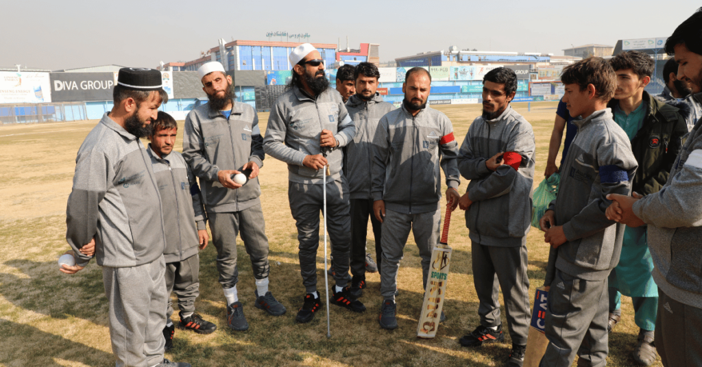 En grupp på tiotals personer står på en cricketplan i matchande, grå overaller. Gräsplanen är torr och himlen är molnfri bakom dem, där byggnader syns. Några av personerna håller i en boll och några håller i racket.