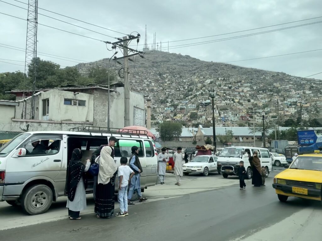 Trafikerad gata i Kabul. I förgrunden syns ett par personer som kliver ur en bil på gatan. I bakgrunden syns ett bergigt landskap täckt av bebyggelse, hus elledningar och biltrafik.