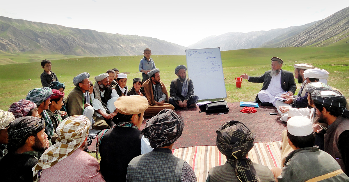 En stor grupp afghanska män med turbaner sitter i en ring på mattor utomhus med bergen i bakgrunden.