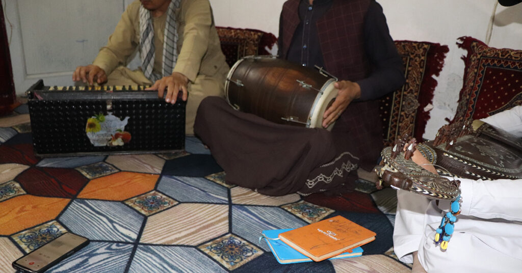 Tre män sitter på en matta och spelar harmonium, tablas (trummor) och rubab (stränginstrument). Deras ansikten syns inte.