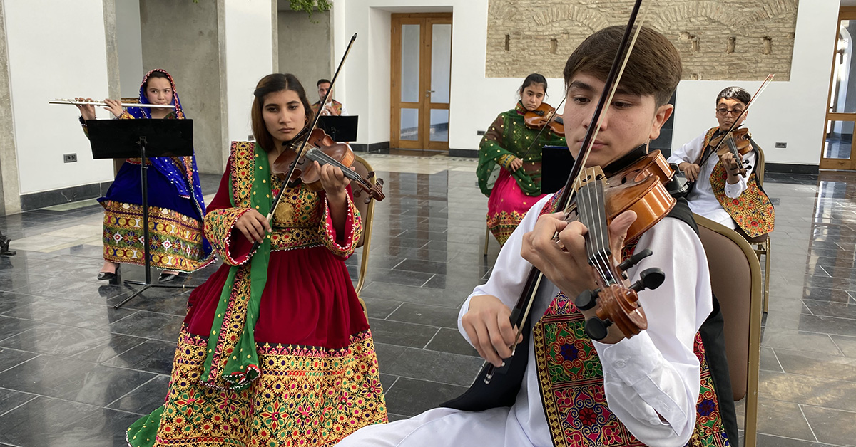 Unga afghaner i traditionella afghanska kläder spelar utspridda i en sal. Närmast bild en ung man som spelar fiol, bakom honom unga kvinnor och män som spelar fiol och tvärflöjt.