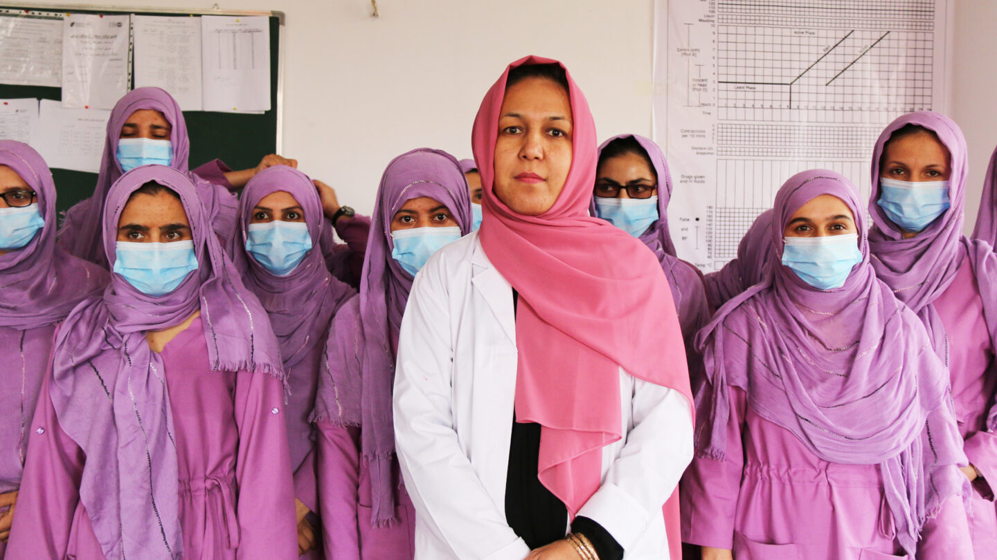 Flera rosa- och lilaklädda kvinnor med hijab, munskydd och professionella arbetskläder står vända mot kameran. Längst fram i mitten står en kvinna i vit rock och rosa hijab utan munskydd.