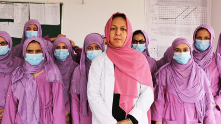 Flera rosa- och lilaklädda kvinnor med hijab, munskydd och professionella arbetskläder står vända mot kameran. Längst fram i mitten står en kvinna i vit rock och rosa hijab utan munskydd.