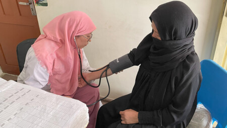 En barnmorska med rosa sjal och vita kläder tar blodtrycket på en kvinnlig patient i svarta kläder och svart sjal i ett undersökningsrum.