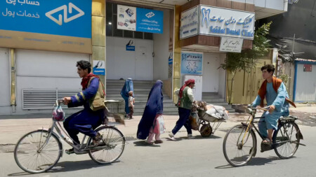 Två män cyklar på en gata i Kabul, kvinnor i burka med barn i bakgrunden.