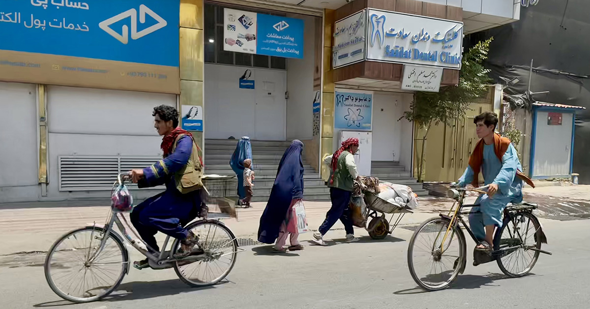 Två män cyklar på en gata i Kabul, kvinnor i burka med barn i bakgrunden.