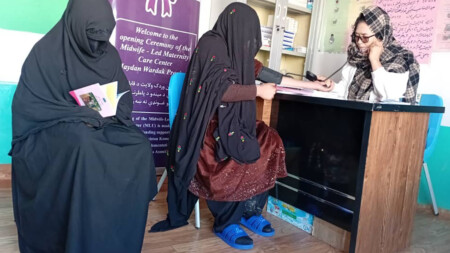 Två kvinnor täckta i burka besöker barnmorskan Marzia på en liten klinik, Marzia sitter till höger bakom ett skrivbord.