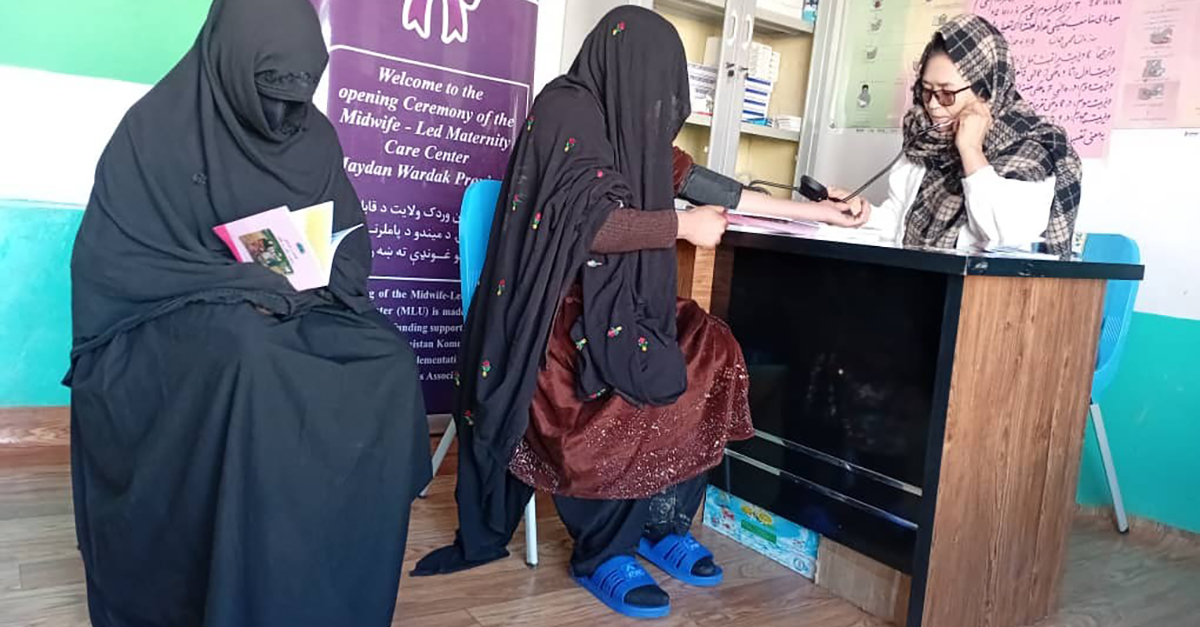 Två kvinnor täckta i burka besöker barnmorskan Marzia på en liten klinik, Marzia sitter till höger bakom ett skrivbord.