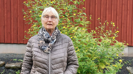 Kvinna står utomhus framför en blomstrande buske och en röd husvägg.