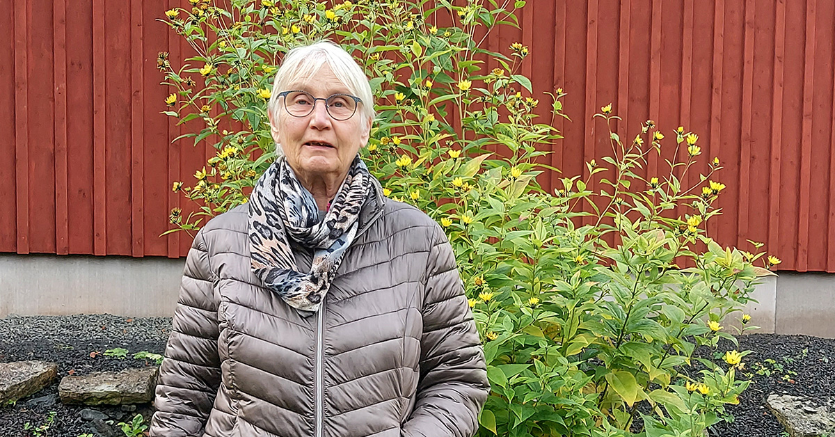 Kvinna står utomhus framför en blomstrande buske och en röd husvägg.