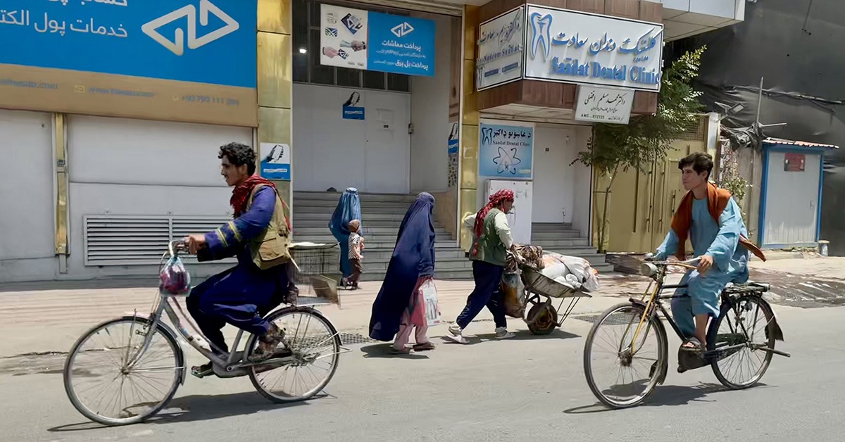 Gatubild från Kabul, med två cyklande män i förgrunden och kvinnor i burka, ett barn och en man med skottkärra i bakgrunden.