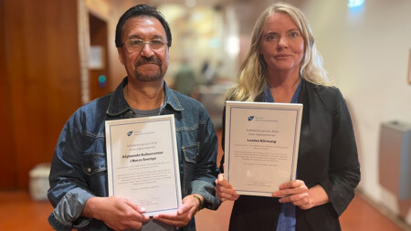 En kvinna och en man står i en korridor med varsitt diplom.