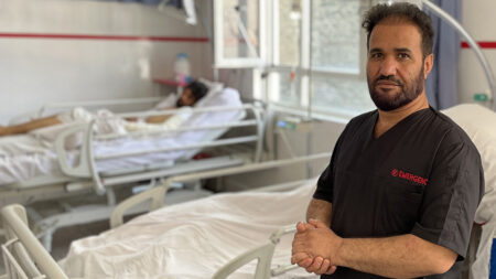 En manlig sjukvårdare i en sjukhussal. I bakgrunden ligger en patient i en säng.