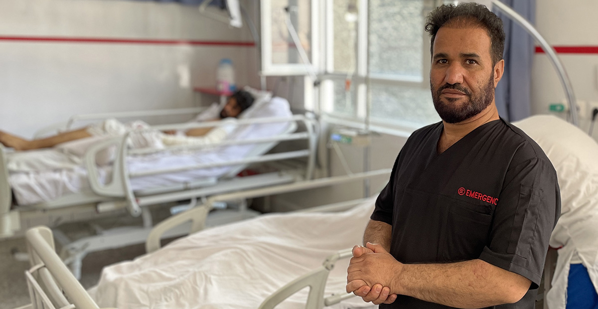 En manlig sjukvårdare i en sjukhussal. I bakgrunden ligger en patient i en säng.